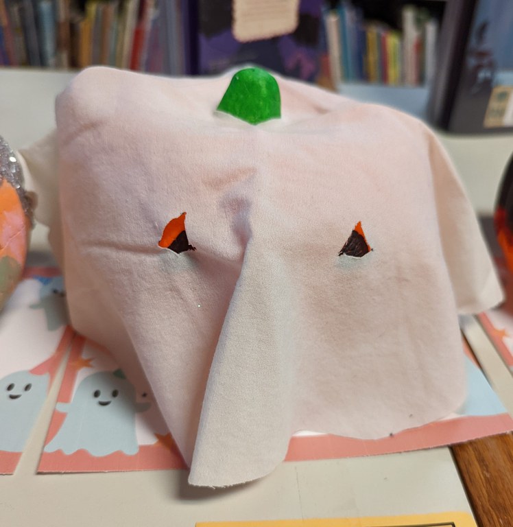 A photo of a small foam pumpkin that has been costumed as a ghost by covering it with a gauzy white piece of fabric. The fabric has cut-outs for the eyes and stem.
