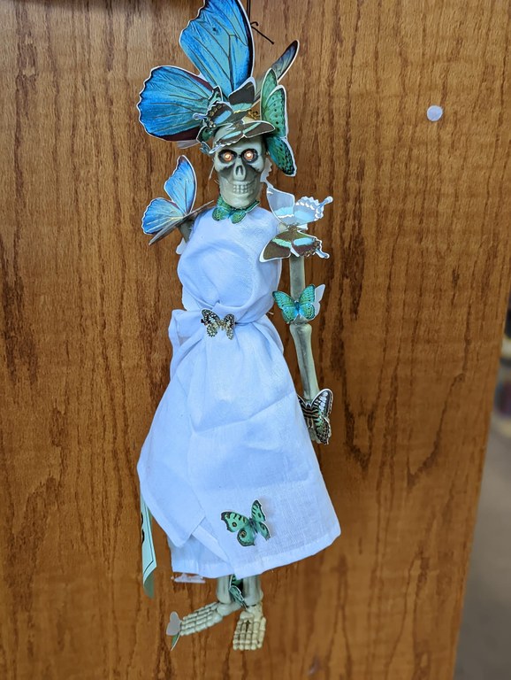 A photo of a plastic skeleton that has been dressed in white and covered in paper butterflies of various sizes. Its eye sockets shine gold.
