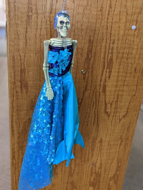 A photo of a small plastic skeleton that has been dressed in a long blue strapless gown, evocative of a mermaid.